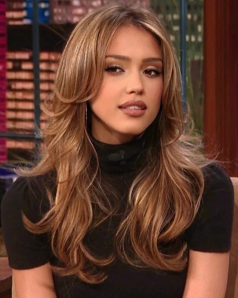 Woman with long honey-blonde hair featuring voluminous waves and side-swept bangs.
