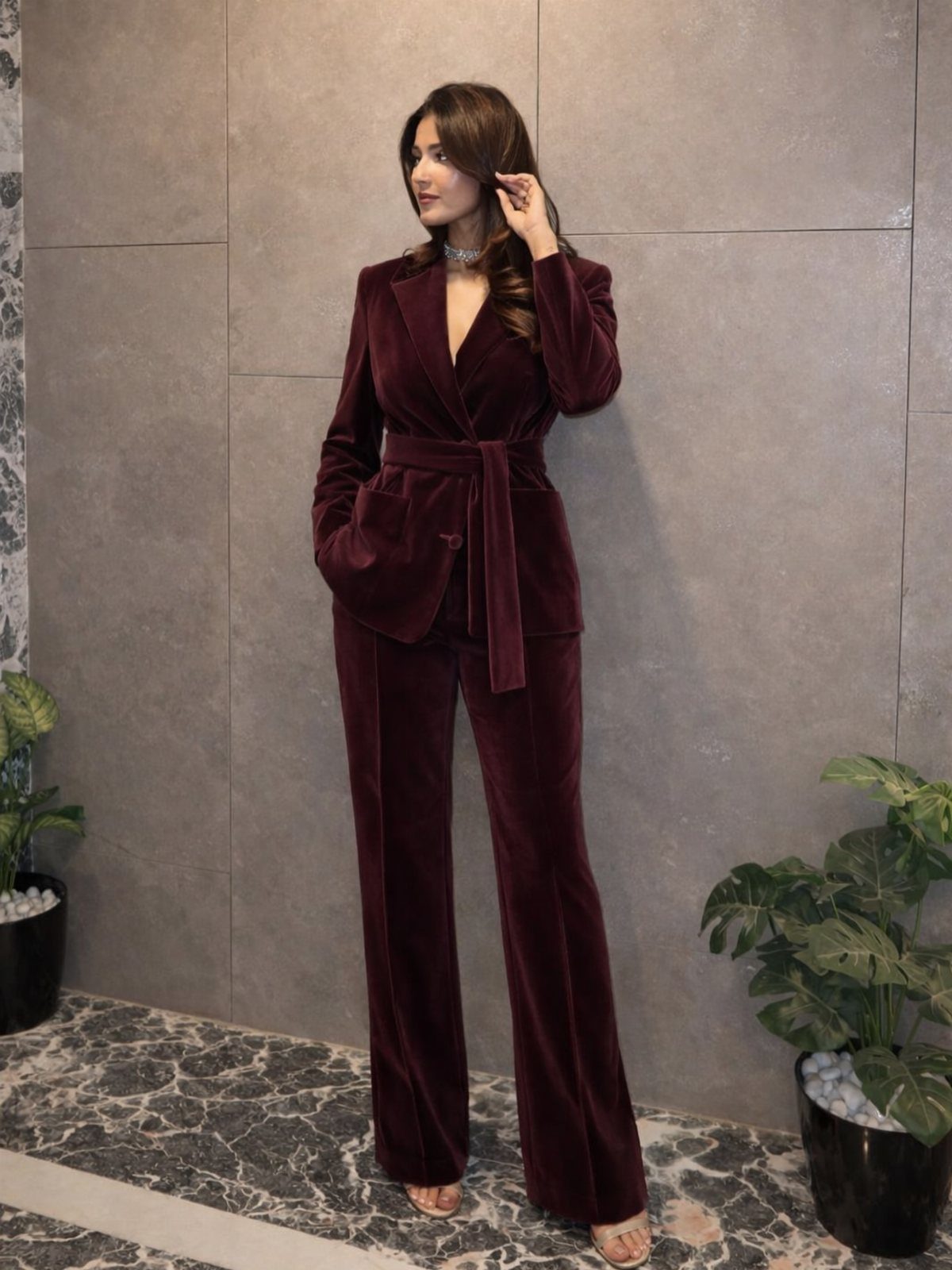 Woman wearing a ruby red velvet blazer and wide-leg trousers against a grey wall.