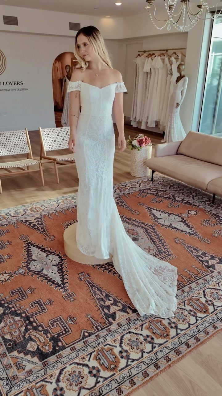Woman in a white off-the-shoulder lace wedding dress standing in a bridal boutique.