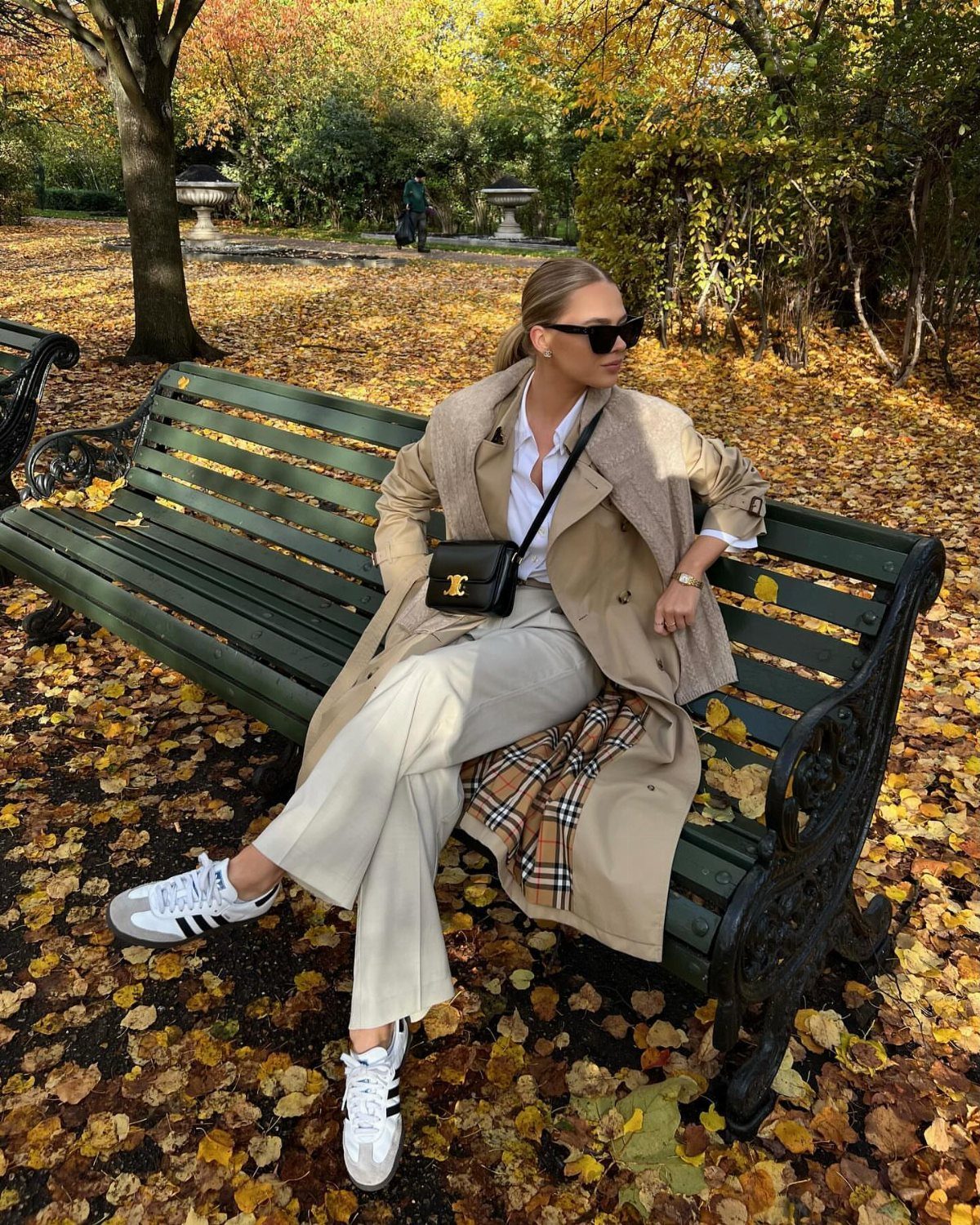 Woman in a beige trench coat and linen trousers sitting on a park bench.