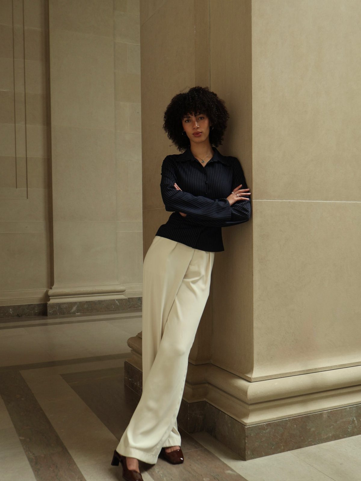 Woman in navy shirt and cream wide-leg trousers leaning against a stone pillar.