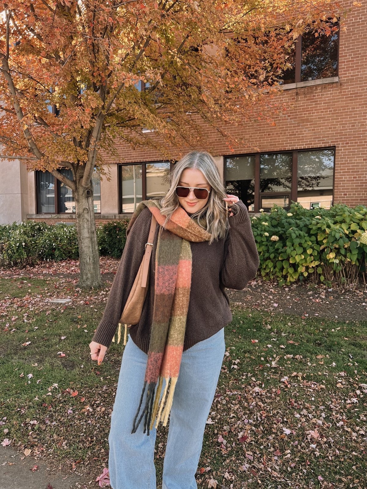 Woman wearing a brown sweater, plaid scarf, and light blue jeans in autumn.