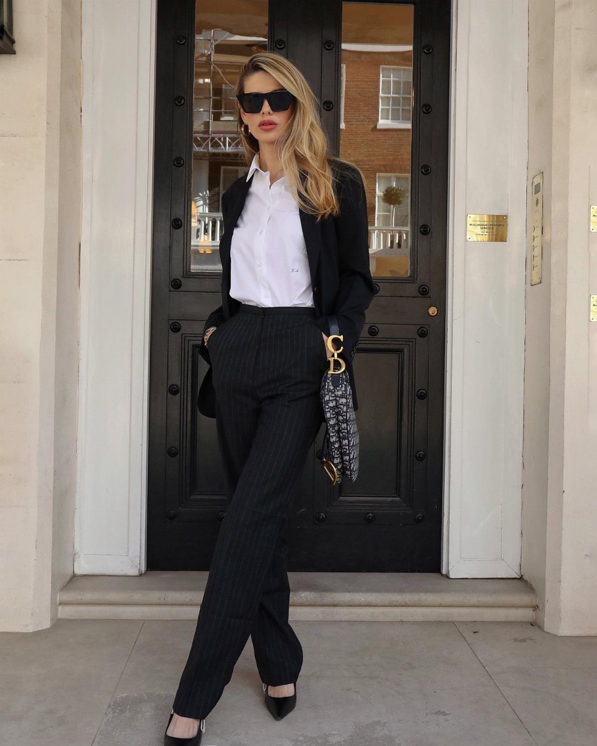 Woman in a black blazer and pinstriped trousers holding a structured patterned handbag.