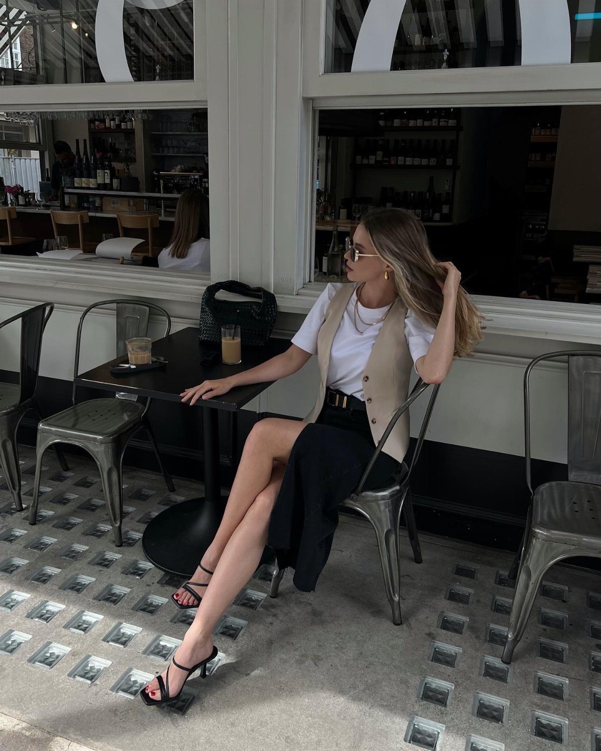 Woman in white t-shirt, beige vest, and long black skirt at a cafe.