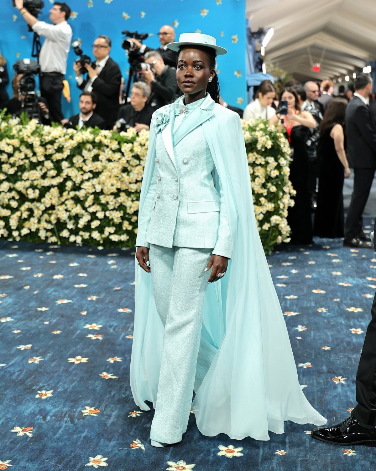 Woman in a light blue suit, hat, and cape at a gala event.