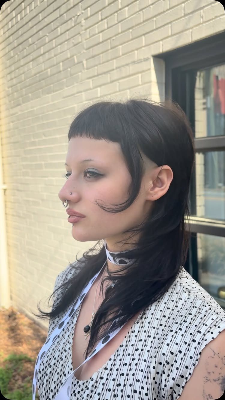 Woman with a dark brown mullet haircut and short straight bangs posing outdoors.