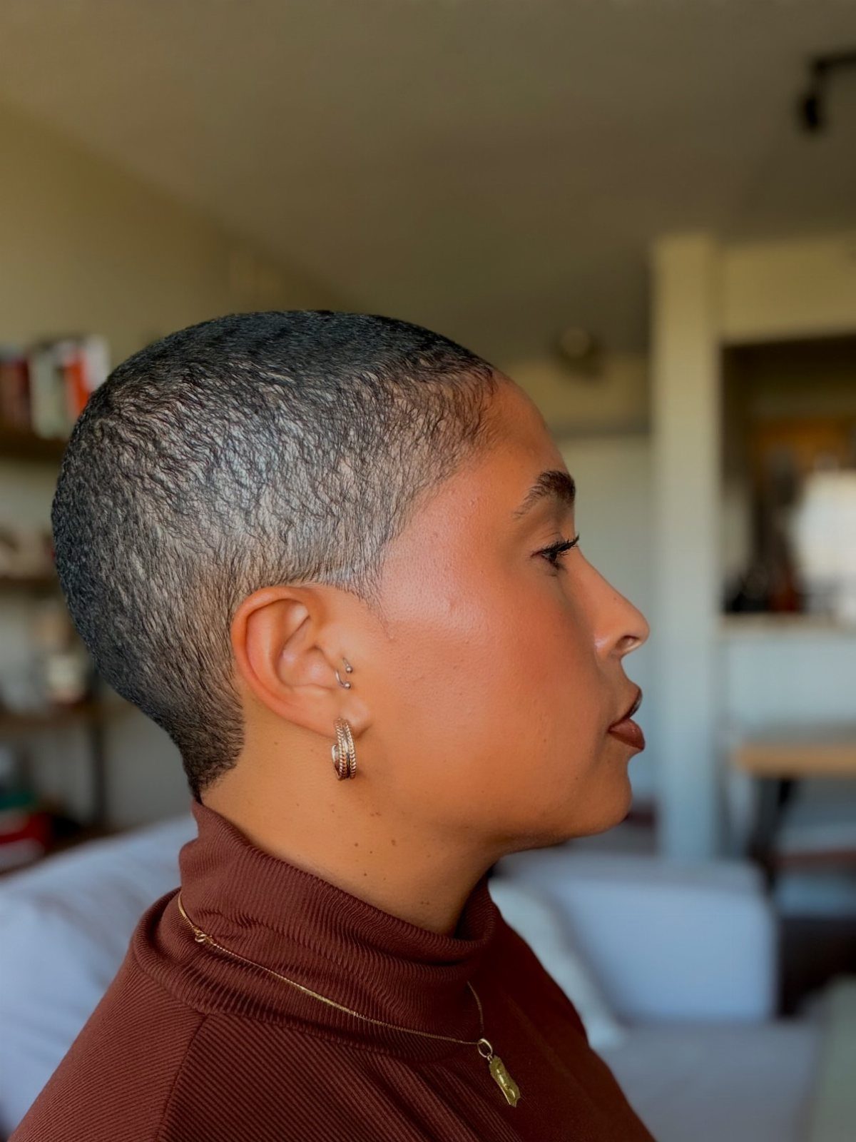 Woman with a short dark buzz cut wearing a brown turtleneck and gold earrings.
