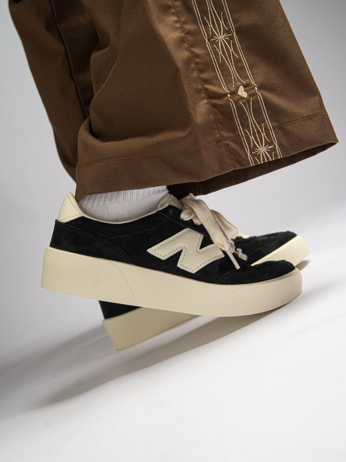 Black New Balance sneakers with cream soles worn with brown trousers and white socks.