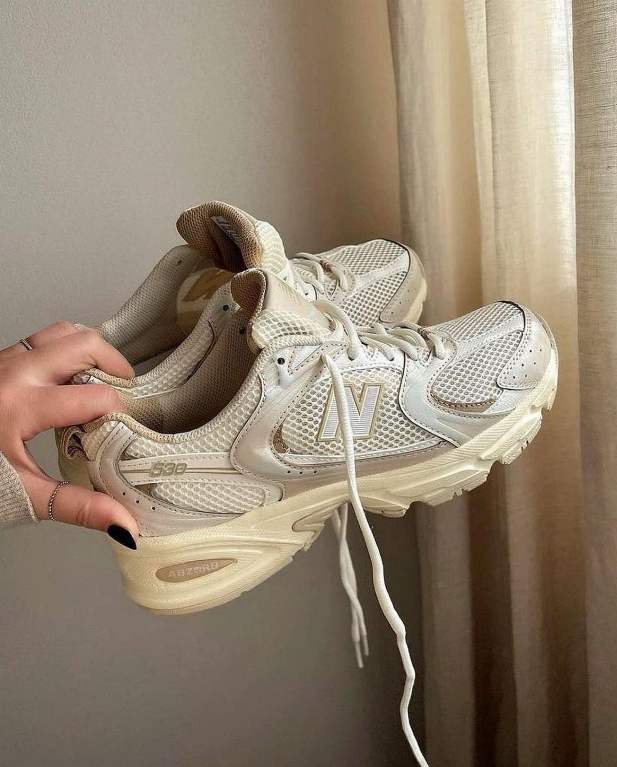 Cream New Balance sneakers held by a hand against a neutral background.