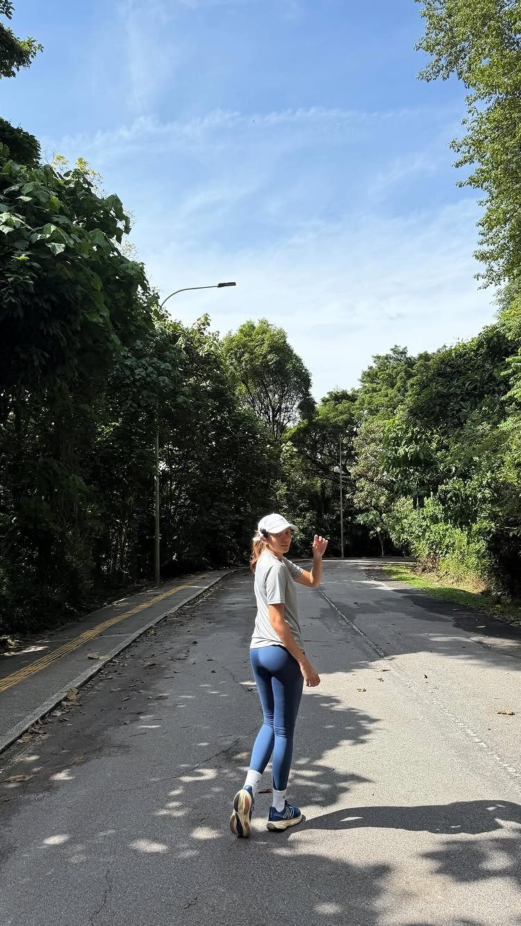 Woman in athletic wear and neutral New Balance sneakers standing on a tree-lined road.