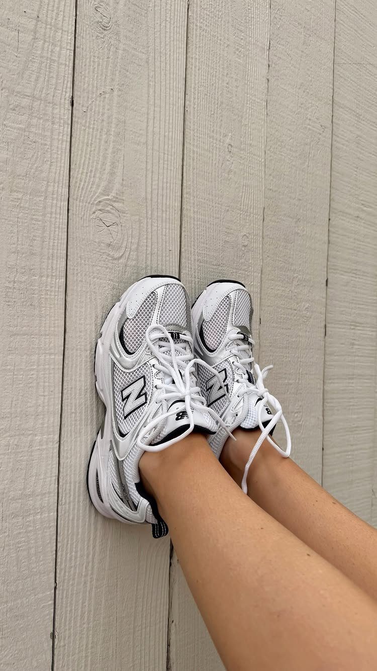 White and silver New Balance 530 sneakers on a light wooden deck floor.
