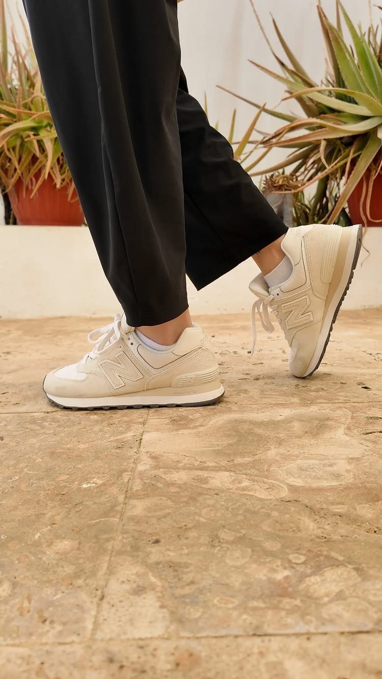 Beige New Balance sneakers worn with black trousers on a stone floor.