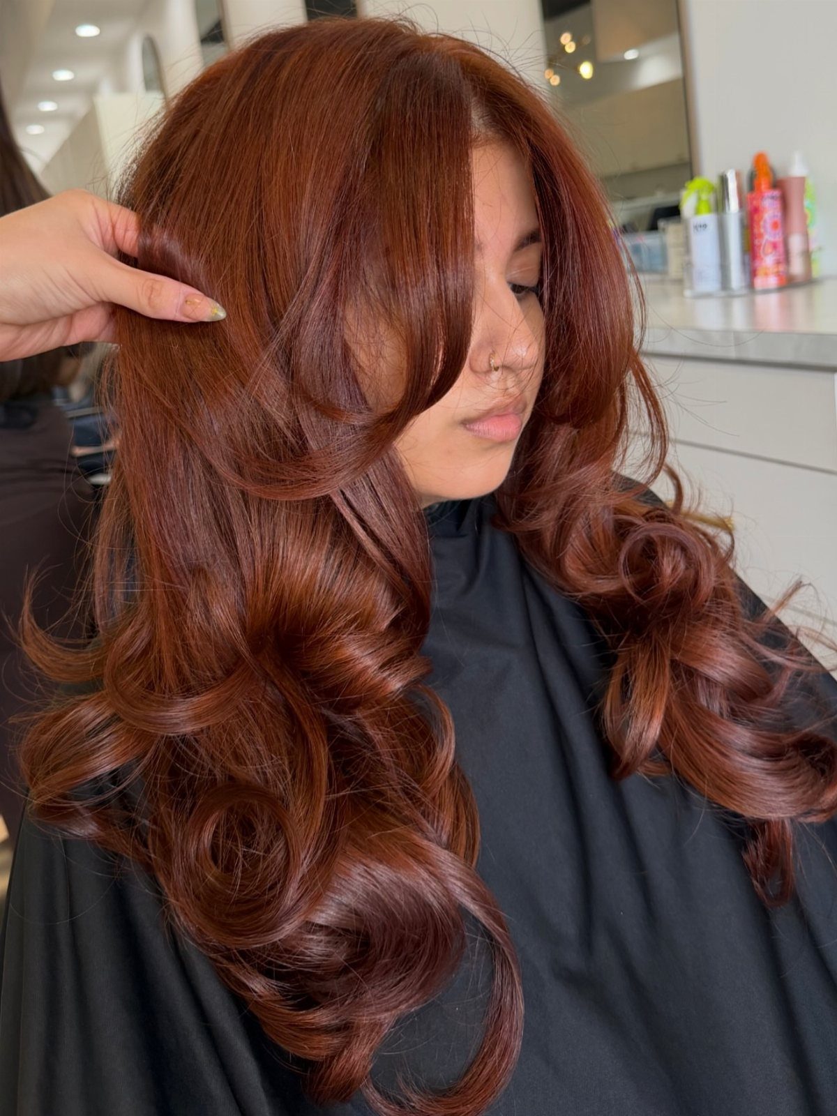 5 No Bleach London Vivid Berry Hair Color Ideas 4 Woman with long, curly, vivid berry-red hair in a bright hair salon setting.