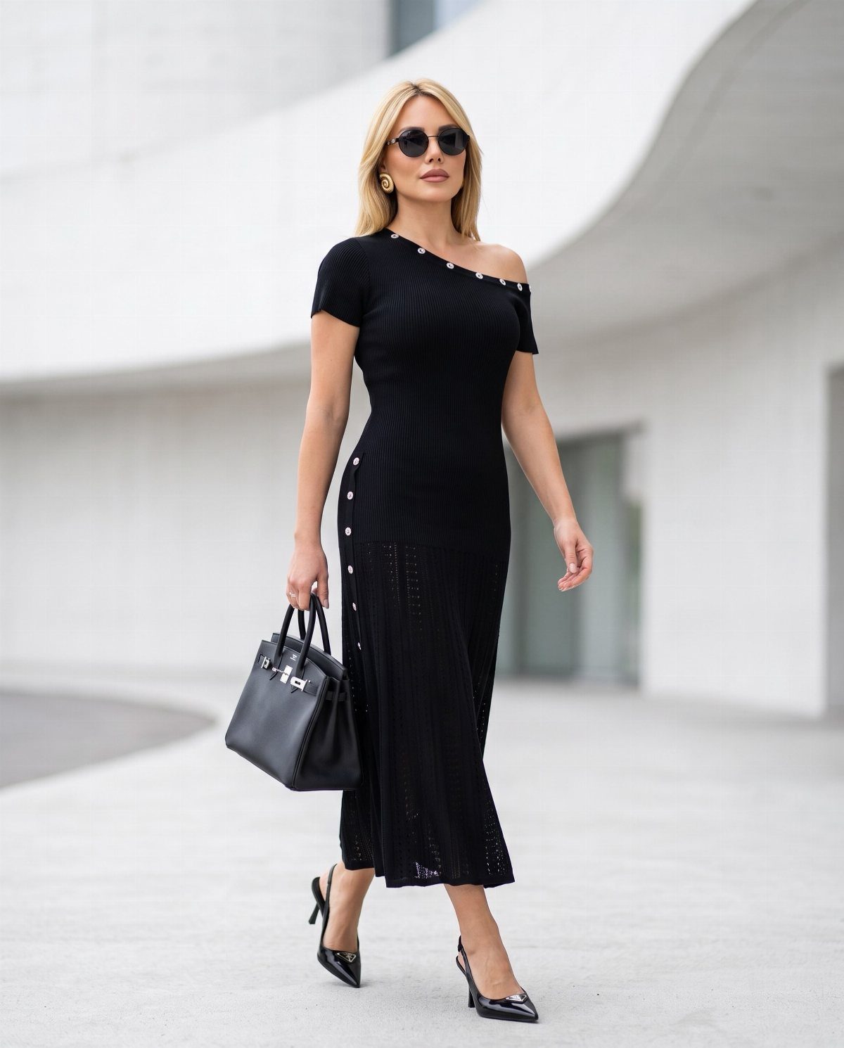 Blonde woman in a black knit midi dress and heels carrying a black handbag.
