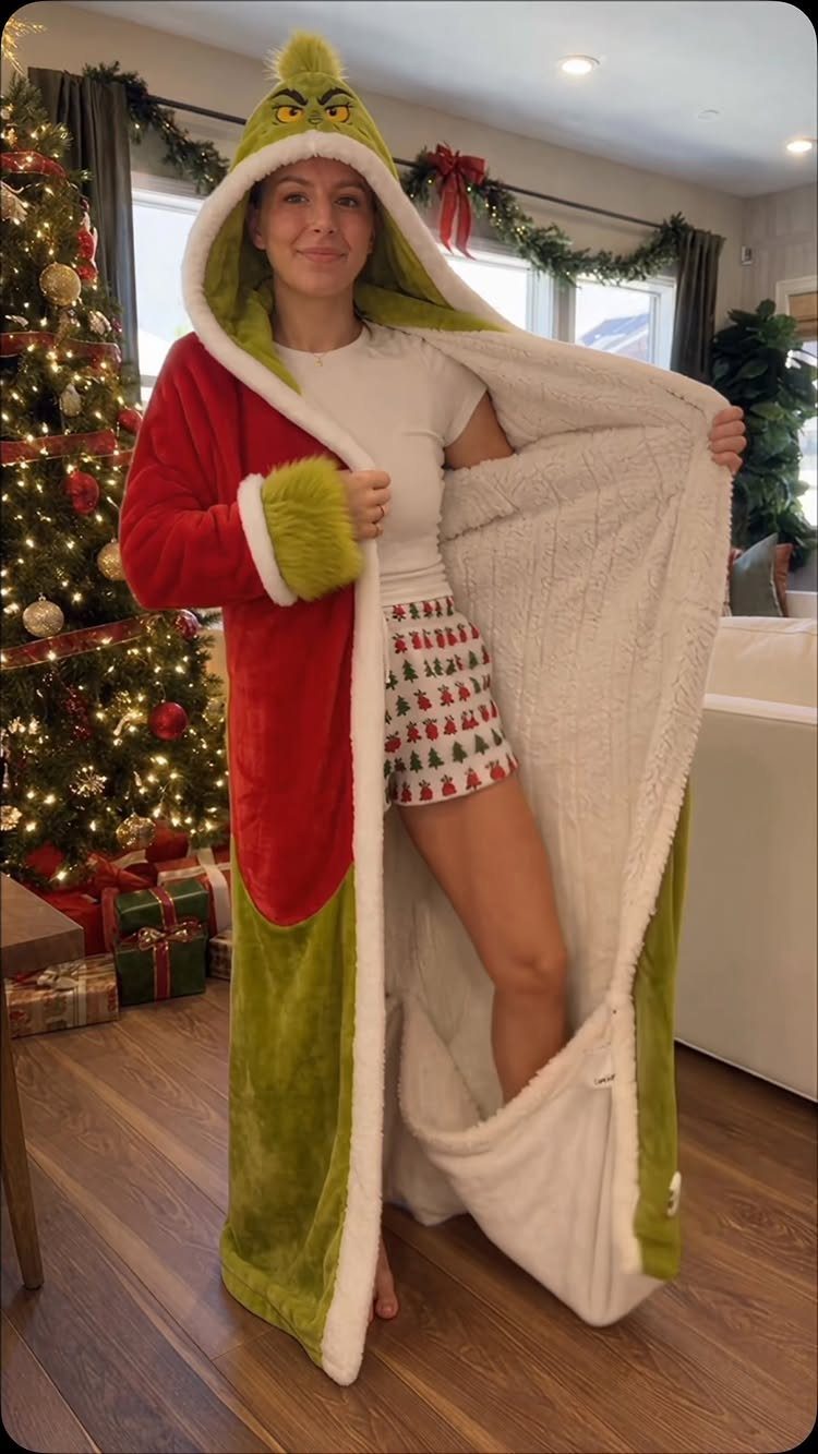 Woman in Grinch robe and Christmas shorts standing by a decorated holiday tree.
