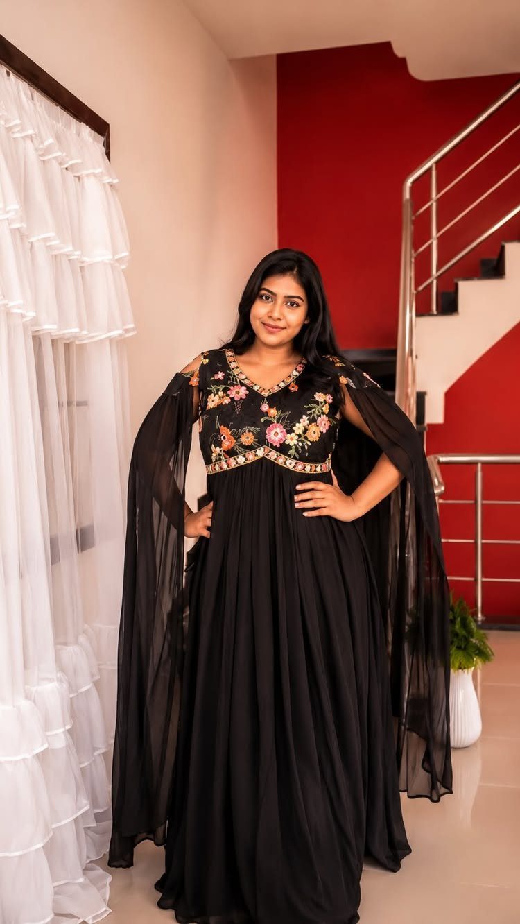 Plus-size woman in a black floral embroidered gown with cape sleeves posing indoors.