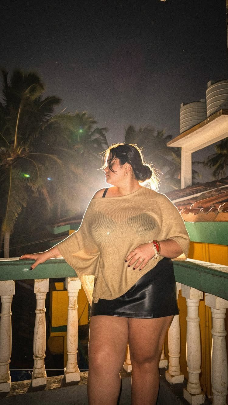 Plus-size woman in a beige off-the-shoulder top and black leather skirt on a balcony.