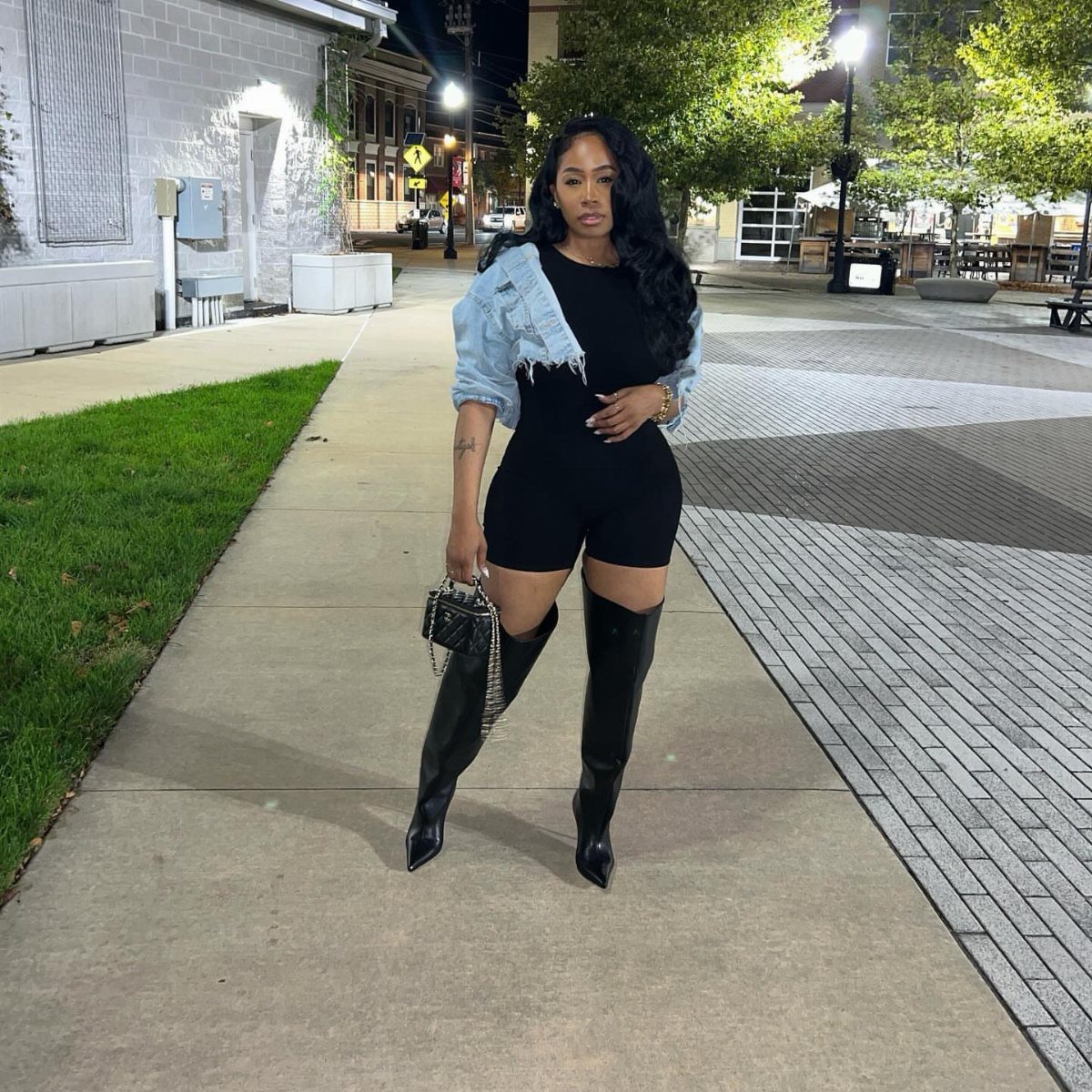 Plus-size woman in a black short jumpsuit, denim jacket, and tall black boots.