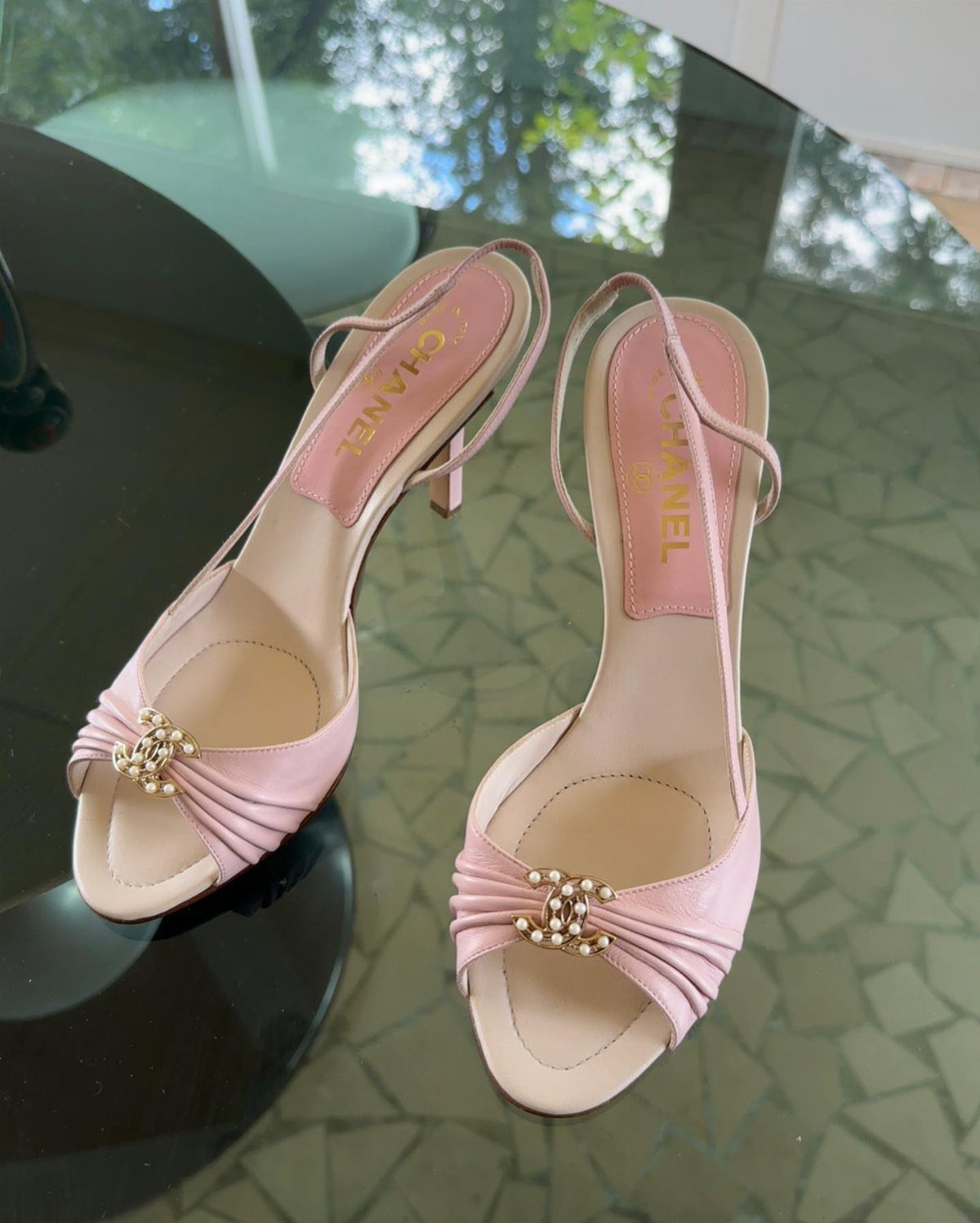 Light pink satin high-heeled sandals with gold logos on a glass table