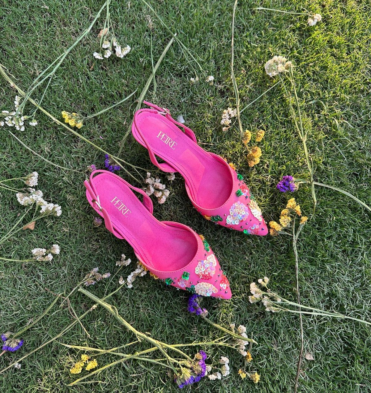 Bright pink pointed-toe heels with floral decorations resting on green grass with wildflowers.