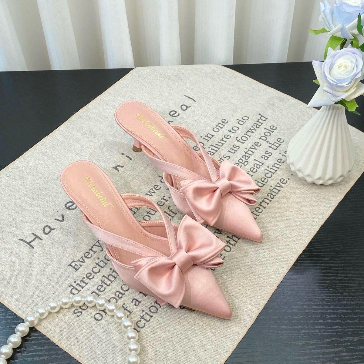 Soft baby pink satin mule heels with large bows on a beige mat.