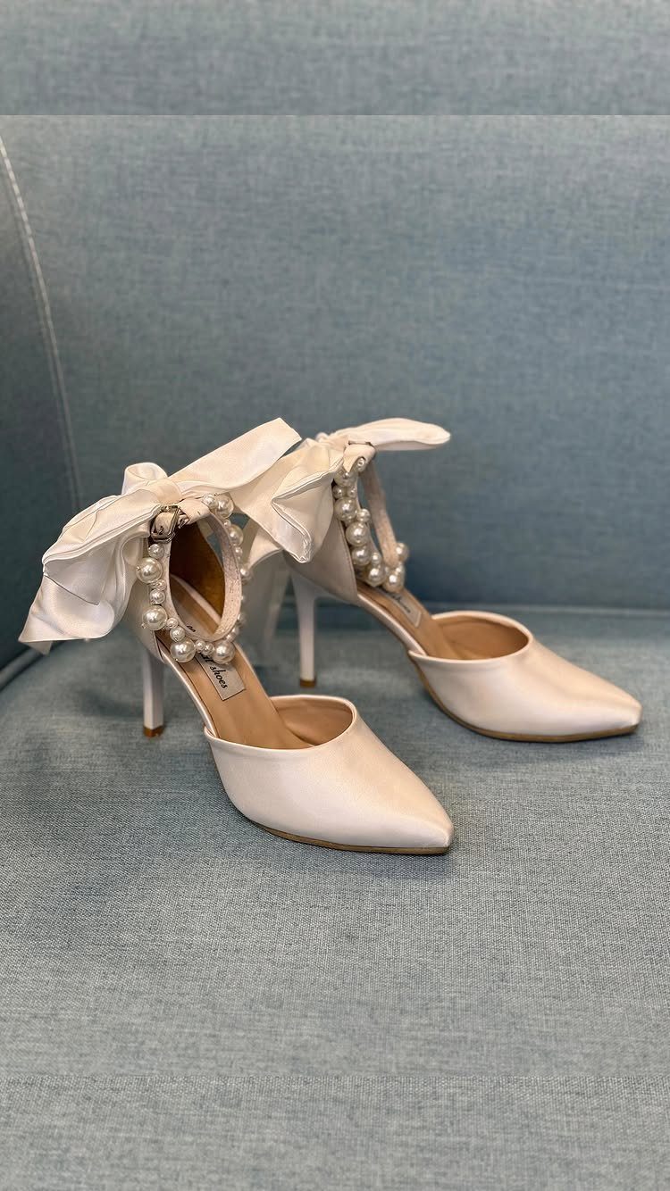 White satin pointed-toe heels with pearl straps and large ankle bows on blue fabric.
