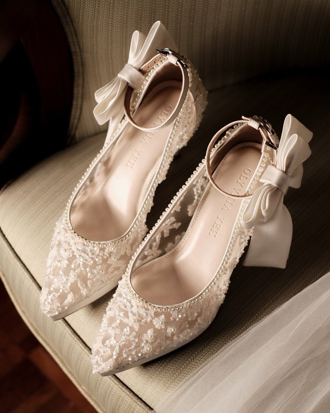 White pointed-toe heels with lace overlays, pearl trim, and satin ankle bows.