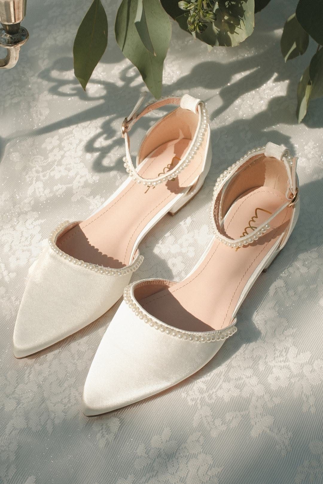 White satin pointed-toe flats with pearl trim and ankle straps on floral fabric.