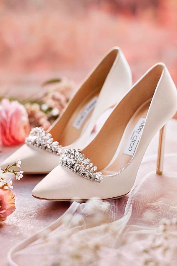White satin pointed-toe heels with crystal crown ornaments and pink floral accents.