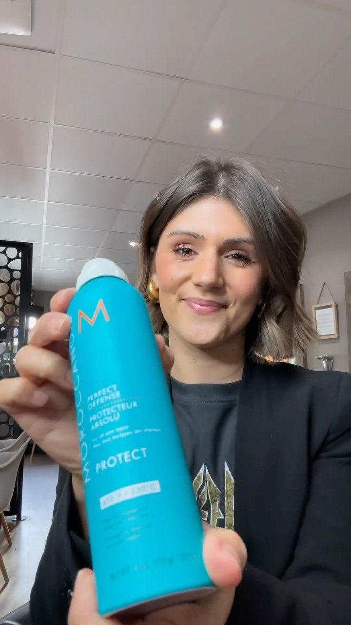 7 Products for Healthy Hair: Scalp Care to Deep Conditioners 4 Woman holding a blue Moroccanoil heat protectant spray bottle in a hair salon.