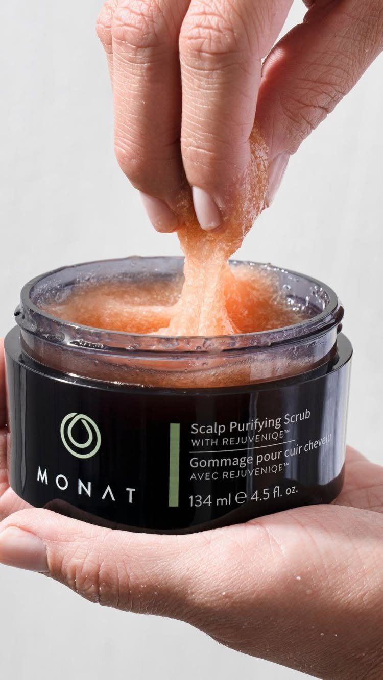 7 Products for Healthy Hair: Scalp Care to Deep Conditioners 1 Hand lifting peach-colored scalp purifying scrub from a black MONAT product jar.