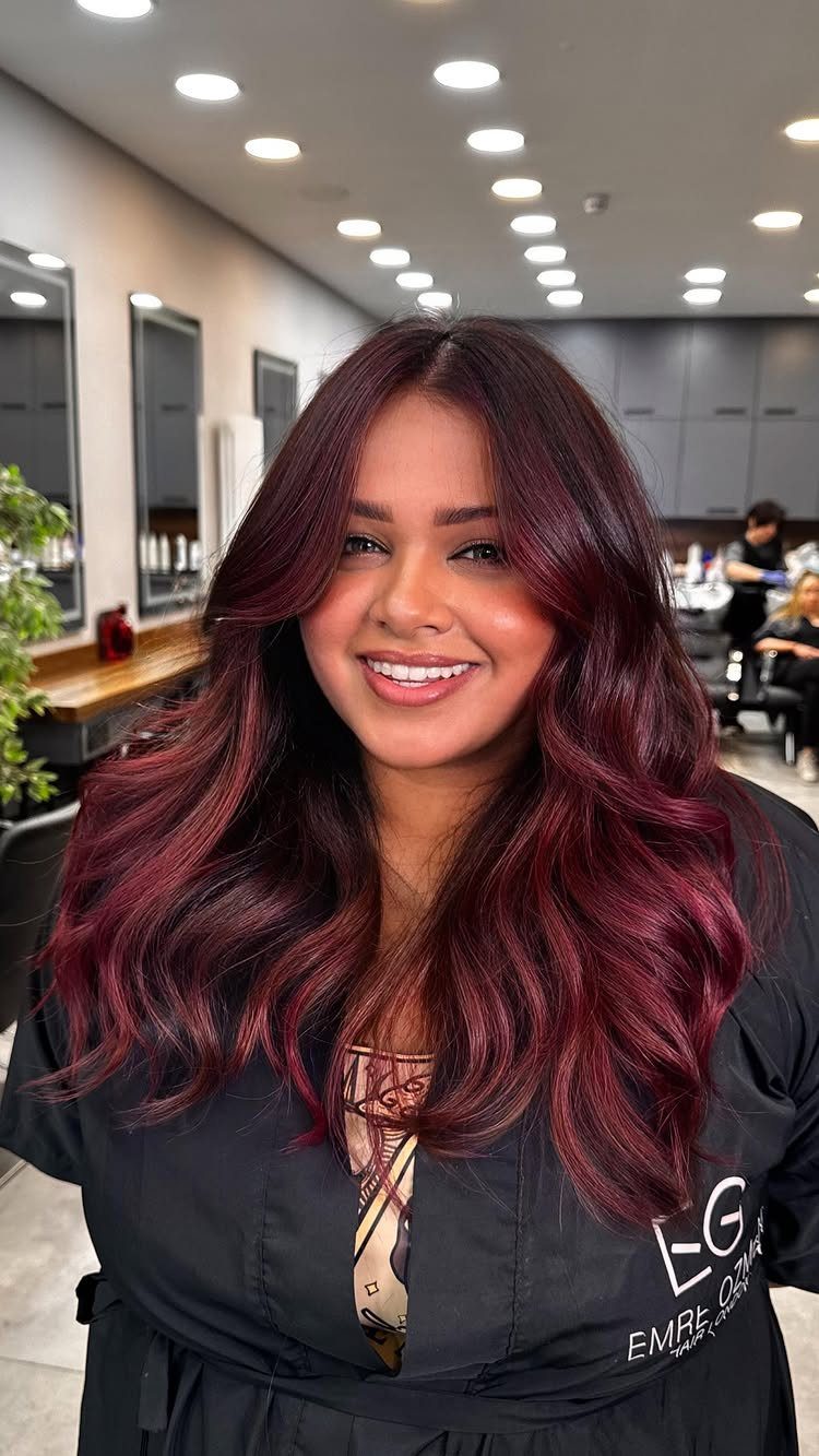 13 Red Undertones Hair Ideas, From Copper to Burgundy 2 Woman with long wavy burgundy hair smiling in a professional hair salon setting
