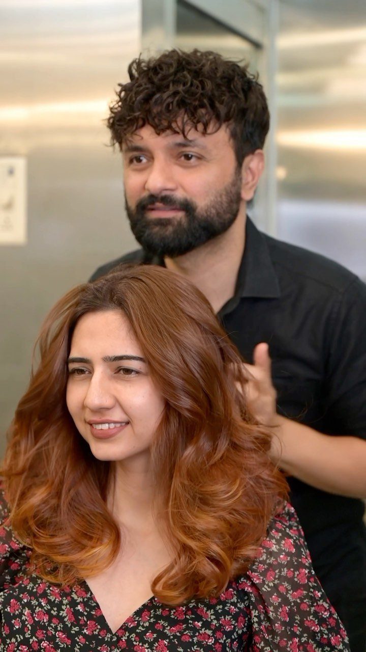 13 Red Undertones Hair Ideas, From Copper to Burgundy 1 Woman with wavy copper-red hair and a man standing behind her indoors.