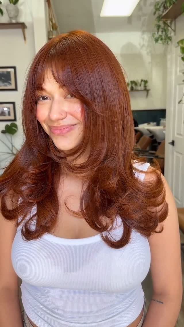 13 Red Undertones Hair Ideas, From Copper to Burgundy 4 Woman with long wavy pumpkin spice reddish-brown hair and bangs smiling indoors.