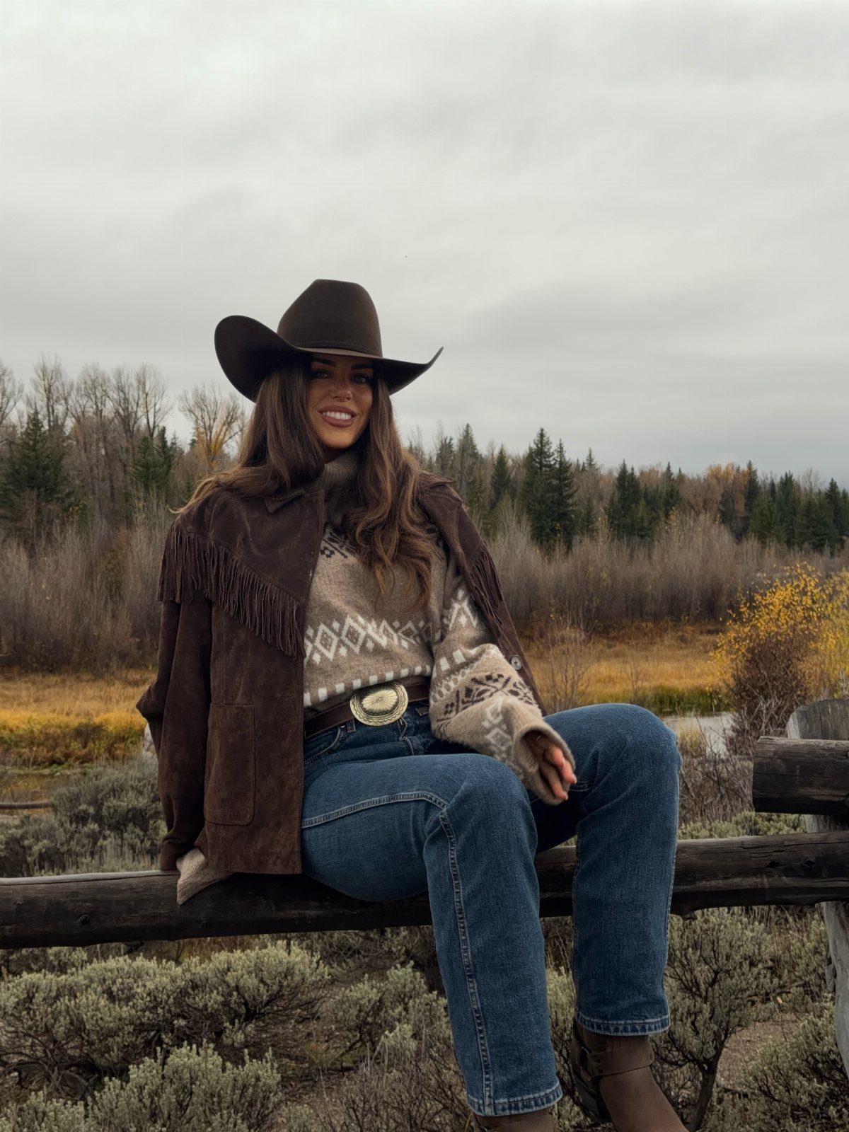 Woman in cowboy hat, fringed suede jacket, and jeans sitting on wooden fence.