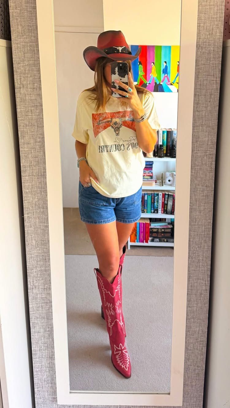 Woman in cowboy hat, graphic tee, denim shorts, and pink cowboy boots.