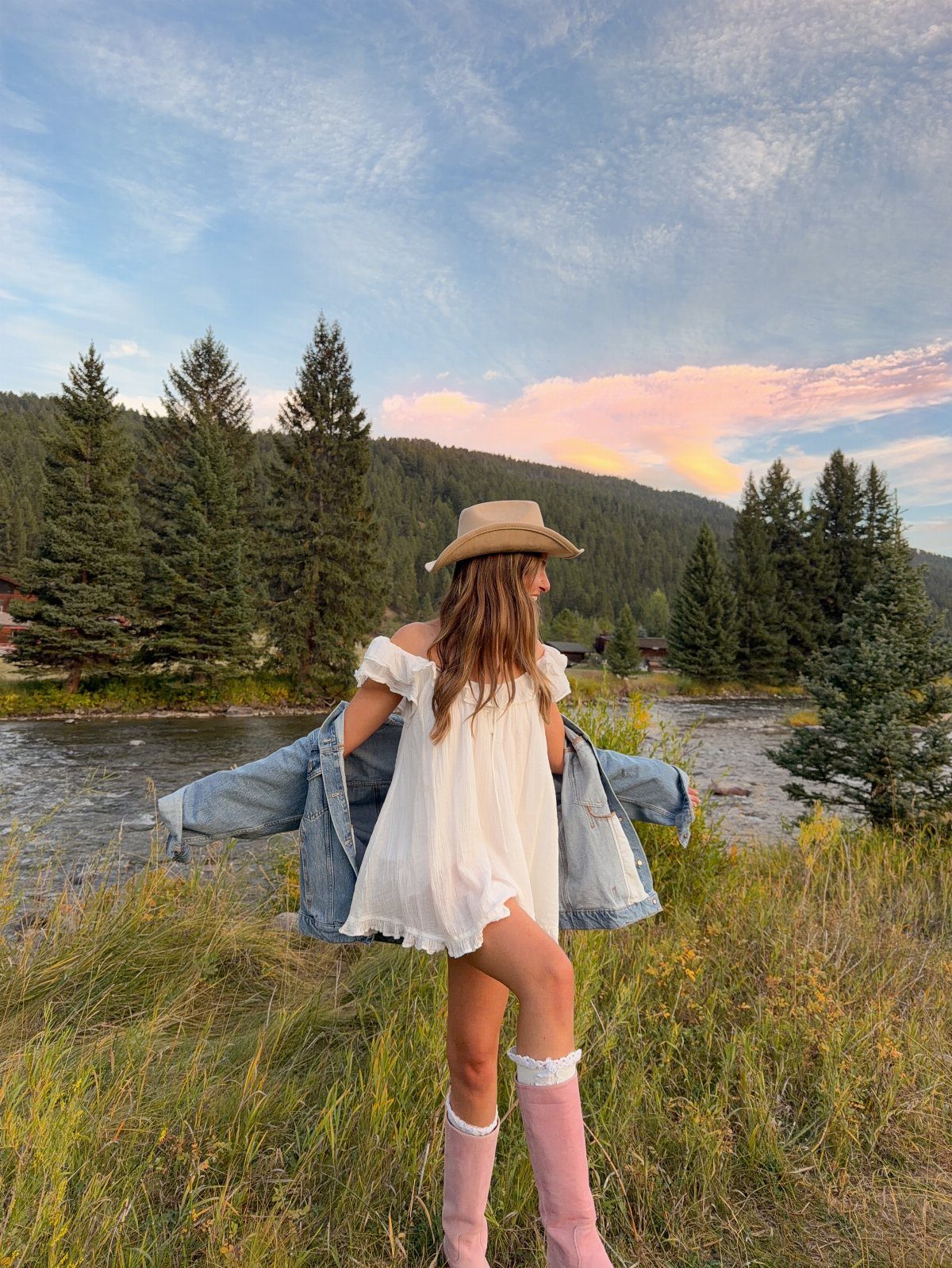 Woman in white mini dress, cowboy hat, and pink boots by a river.