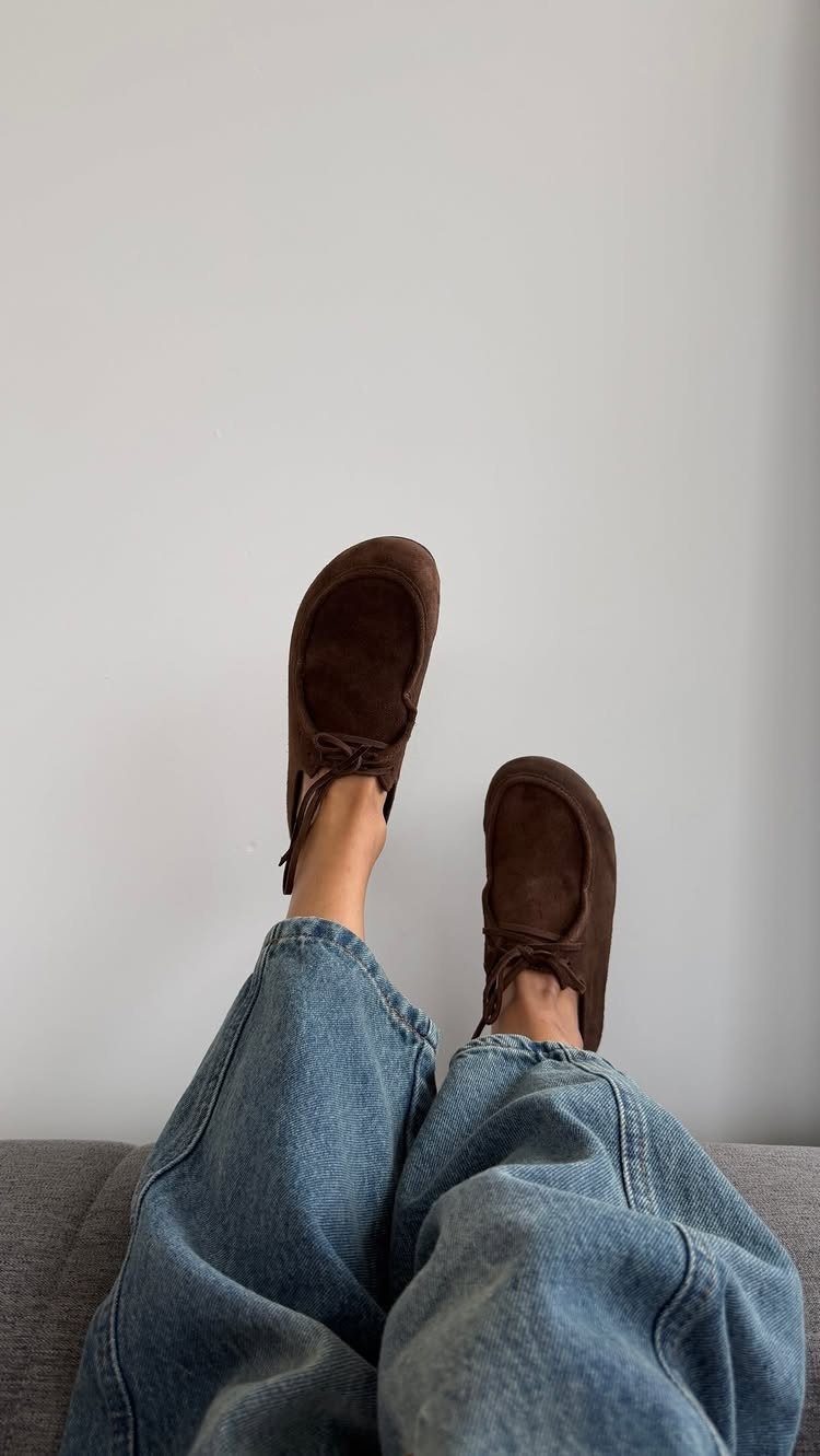 Brown suede mules worn with blue flared denim jeans against a white wall.