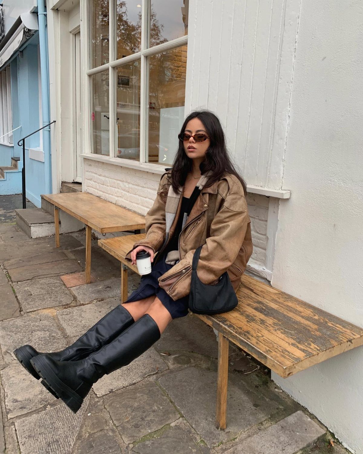 Woman in tan leather jacket and black slouch boots sitting on wooden bench