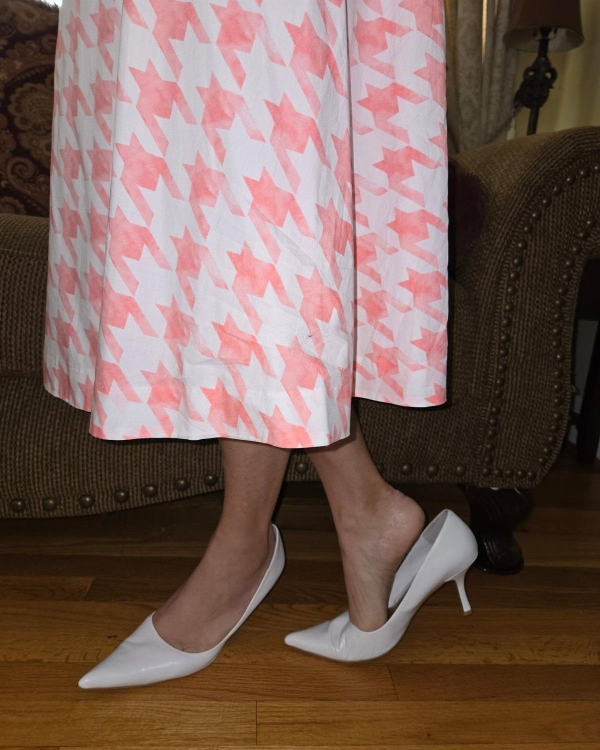 White pointed-toe pumps worn with a pink and white patterned skirt on wooden flooring.