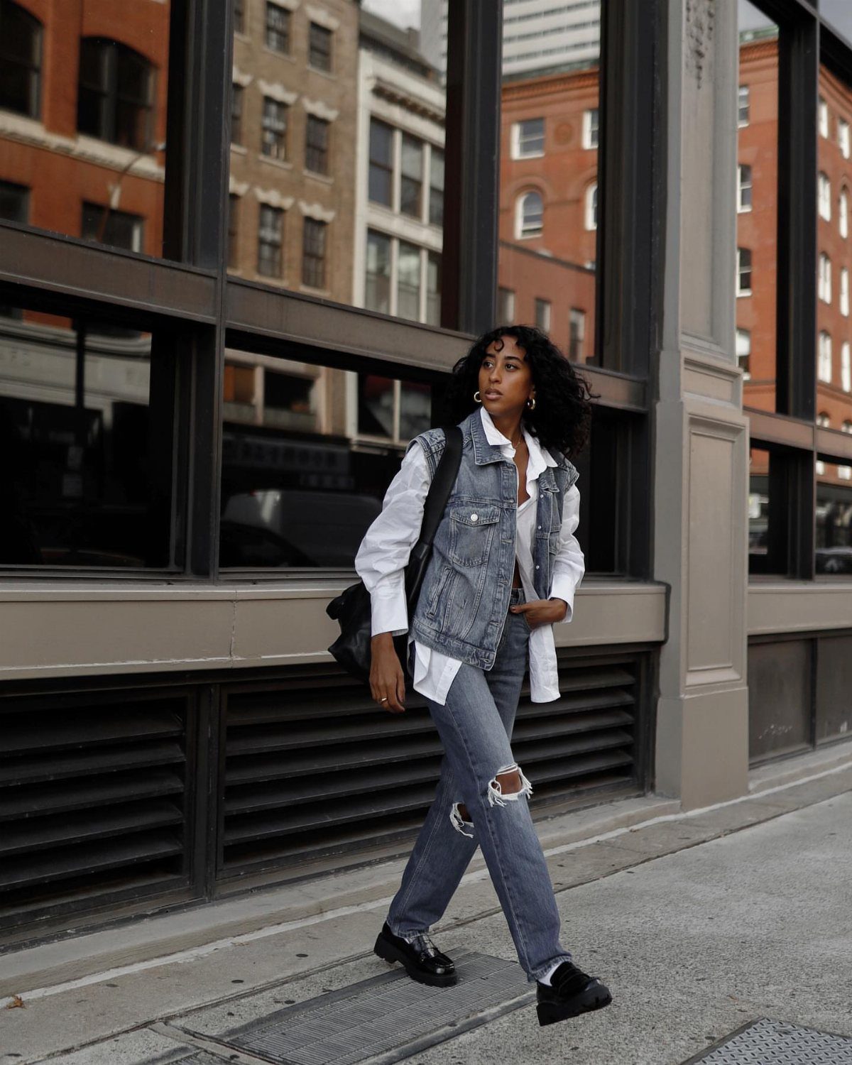 Woman in white shirt, denim vest, and ripped jeans walking on city street