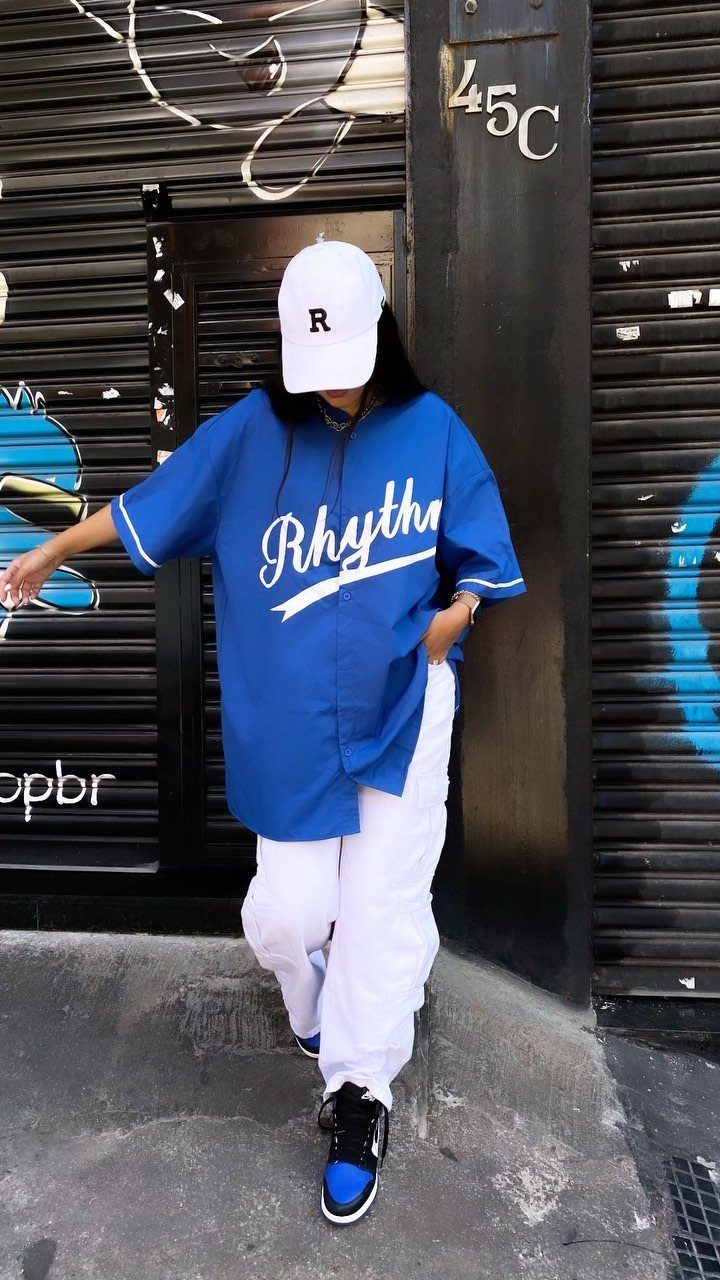 Woman in oversized blue baseball jersey and baggy white pants posing by black shutter.