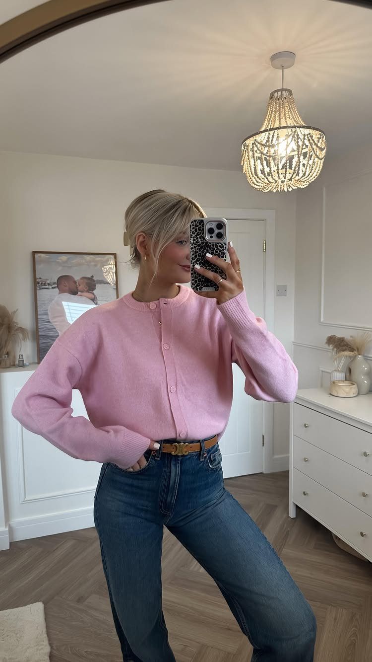 Woman wearing a pink cardigan and blue jeans taking a mirror selfie indoors.
