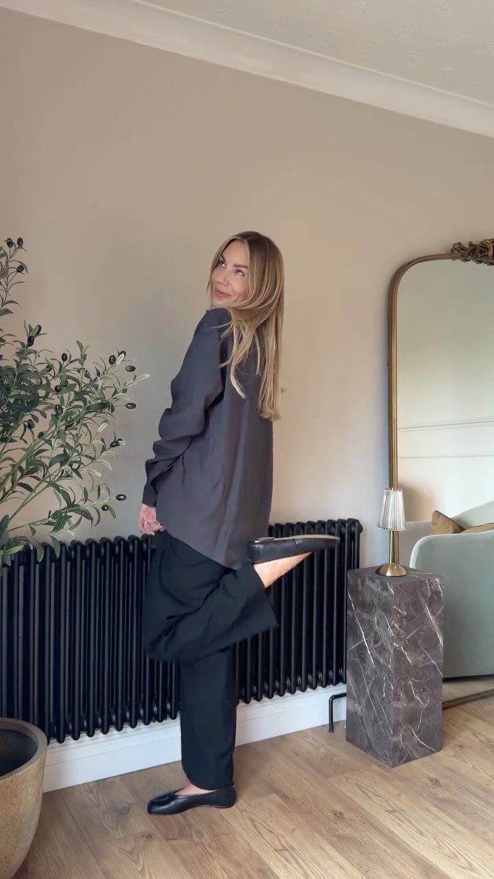 Woman in grey shirt and black trousers wearing black classic loafers indoors.