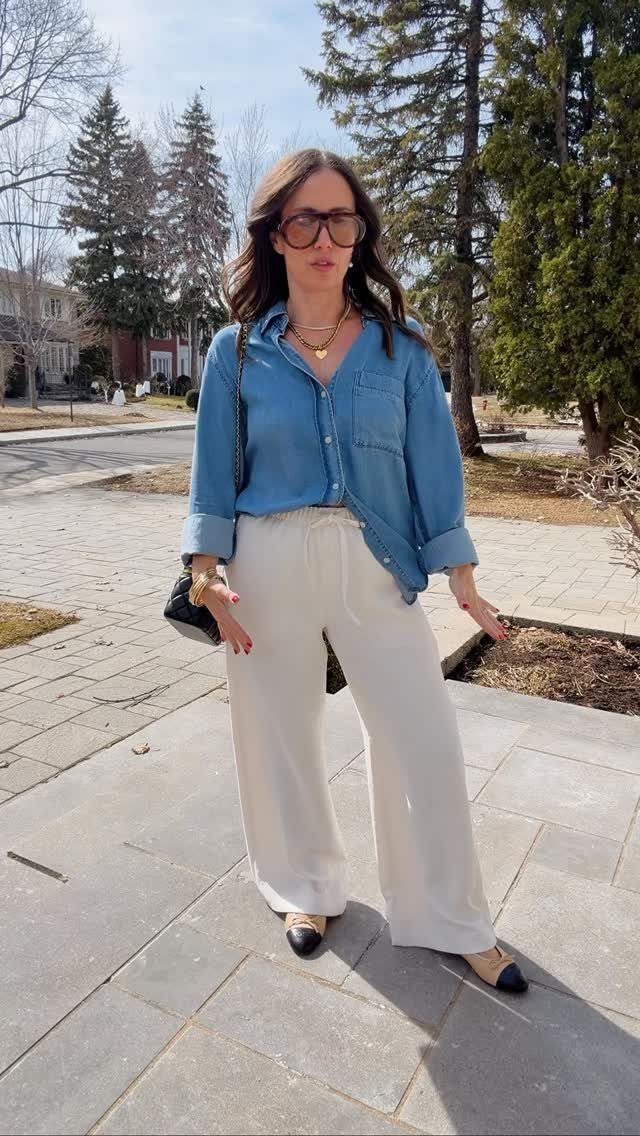 Woman wearing a blue denim shirt and wide-leg white trousers on a sunny walkway.