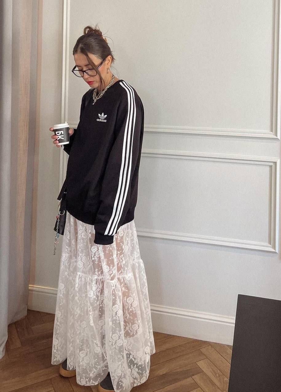 Woman wearing a black Adidas tracksuit jacket and a long white lace skirt.