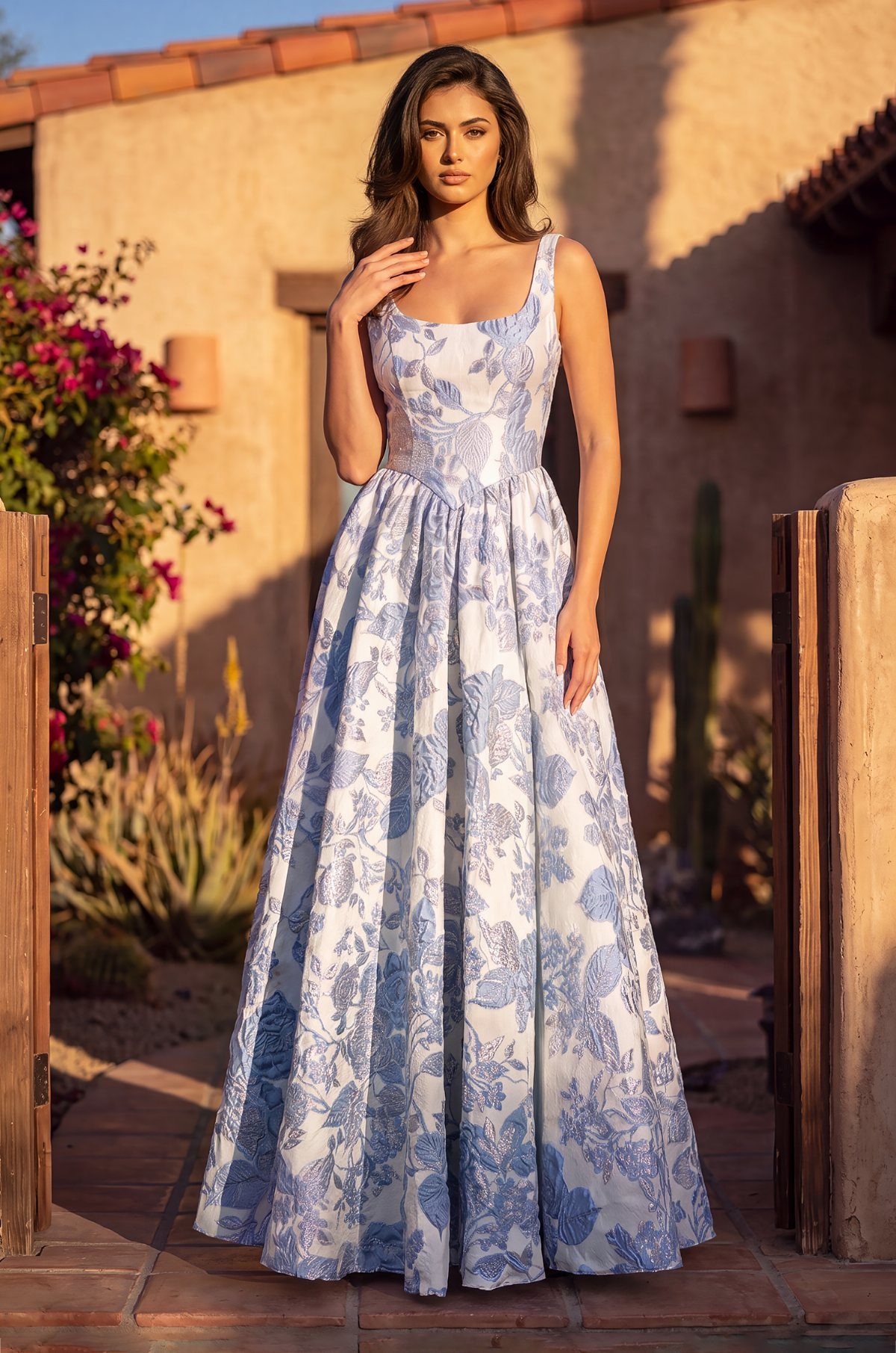 Woman wearing a light blue floral brocade A-line dress in a sunny courtyard.