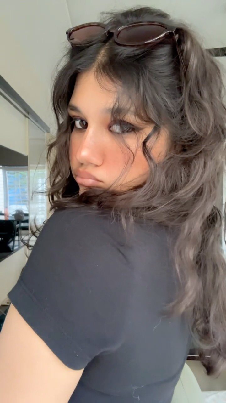 Woman with long dark wavy hair and sunglasses on her head posing indoors.
