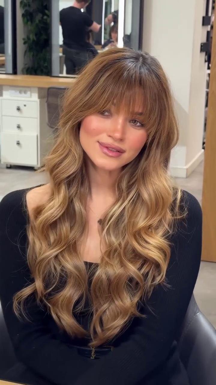 Woman with long honey-blonde layered wavy hair and face-framing curtain bangs.