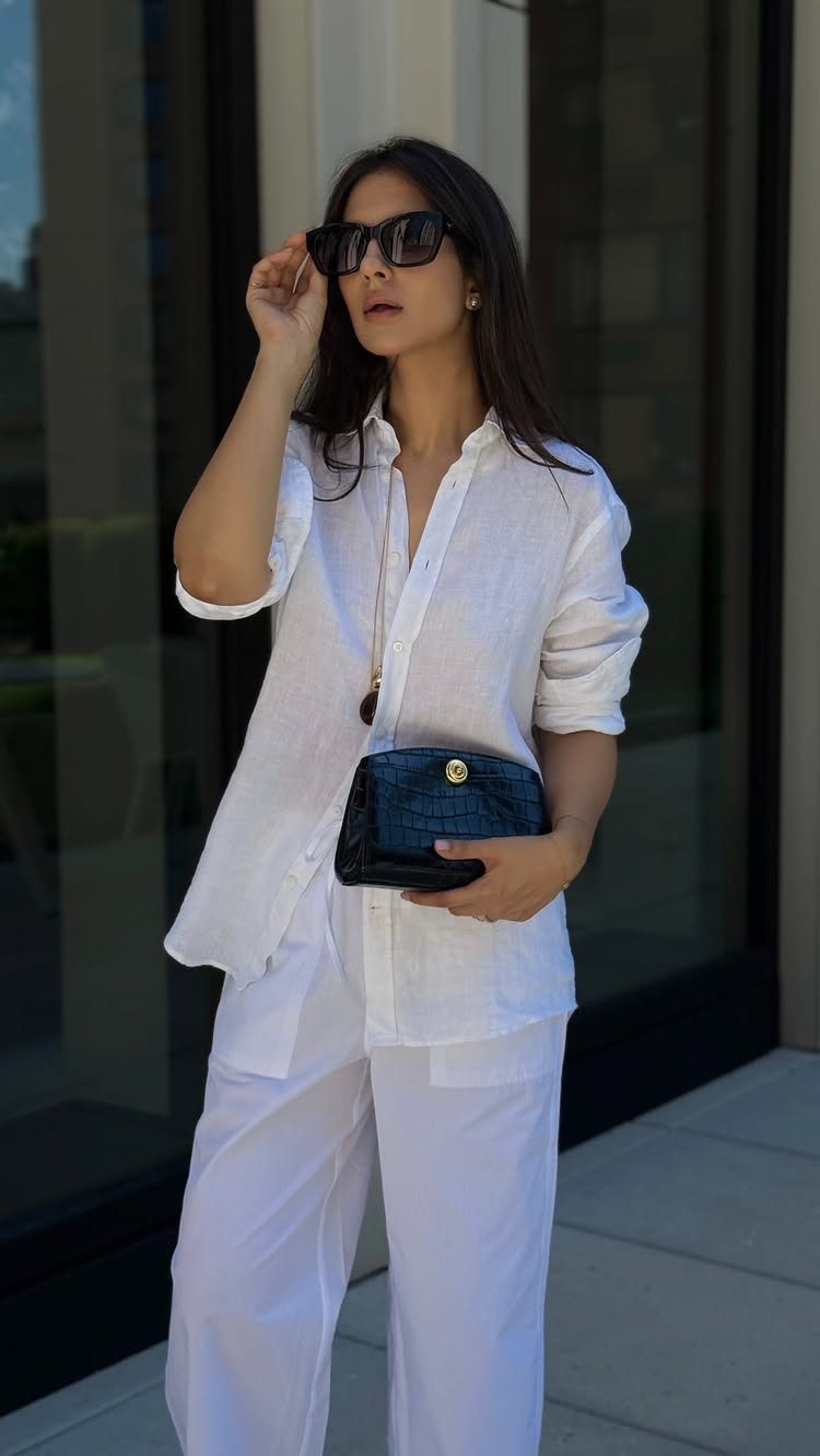 Woman in a white button-down shirt and white linen trousers holding a black clutch.